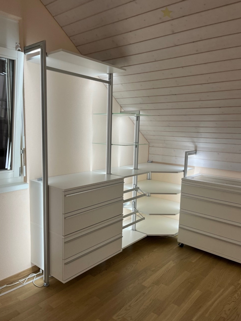 Kundenfoto begehbares Schranksystem unter Dachschräge in Weiß Kundenfoto begehbares Schranksystem unter Dachschräge mit weißen Schubladenelementen und Glasböden – maßgefertigte Stauraumlösung